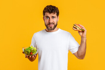 Confused Guy Holding Burger And Vegetable Salad Choosing Diet, Studio