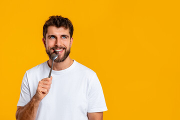 Hungry guy holding and biting spoon looking aside, yellow background