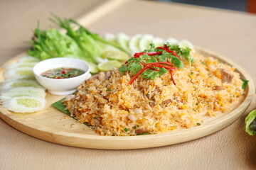 Pork fried rice with egg topped with coriander leaves and sliced ​​red chili. Fried rice served on a wooden plate decorated with sliced ​​cucumbers, lime wedges, spring onions and chili fish sauce.
