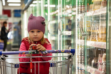 Pensive little girl shopper 5 year old in red clothes get buy in food store, thought looking at camera. Kid girl sit in shopping cart buying in supermarket. Retail purchase concept. Copy ad text space