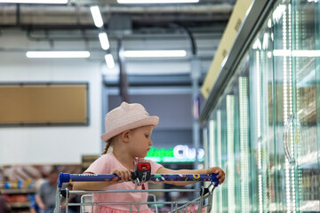 Child little girl sitting in shopping cart near shop windows in food store, pensive looking away....