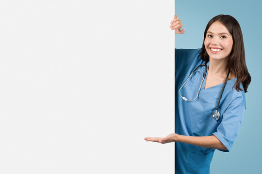 Young nurse in blue, pointing at empty board, blue backdrop - Powered by Adobe