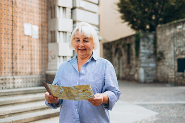 Mature blonde woman tourist looking at city map at street. Cheerful 60s woman traveling abroad in summer. Travel and active lifestyle concept