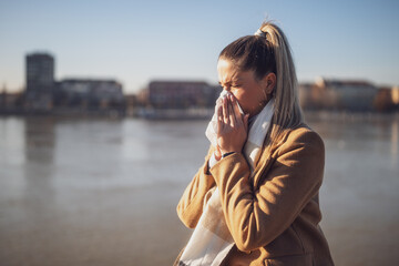 Woman blowing nose while sitting by the river on a sunny winter day.	Toned image. - obrazy, fototapety, plakaty