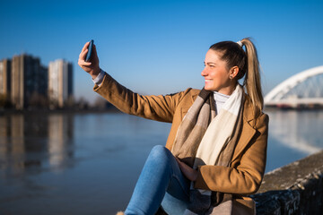 Beautiful woman in warm clothing taking selfie with phone and enjoys resting by the river on a sunny winter day.