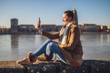 Beautiful woman in warm clothing taking selfie with phone and enjoys resting by the river on a...