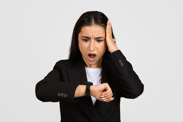 Shocked businesswoman in suit, looking at clock with open mouth and touching head, isolated on gray studio background