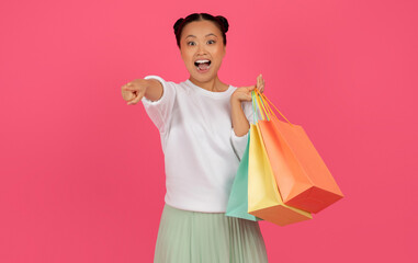 Cheerful young asian lady with shopping bags pointing at camera
