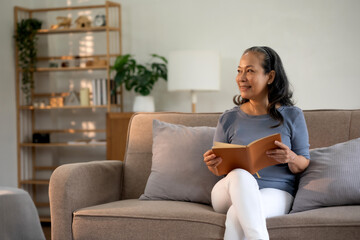 Obraz premium Senior asian old woman sitting on sofa in the living room reading a book to relax on a leisure day.