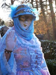 Personne portant costume et masque sur le thème du carnaval de Venise, avec chapeau et foulard dans les tons bleu-blanc, levant la main pour saluer le public
