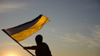 Young man in military uniform waving flag of Ukraine against beautiful sunset at background. Male ukrainian army soldier lifted national banner at countryside. Victory against aggression