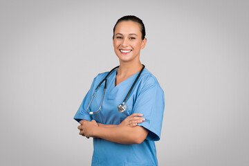 Happy nurse lady with folded arms posing wearing blue workwear and standing on gray studio background, smiling to camera