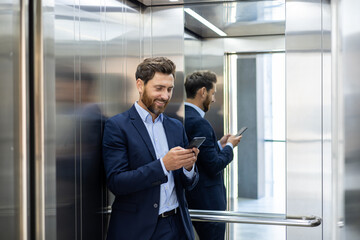 Smiling businessman in suit using smartphone in modern elevator, reflecting success and connectivity © Tetiana
