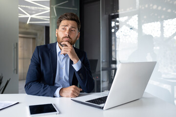 Thoughtful businessman in office pondering strategy with laptop and tablet, concept of planning and decision making