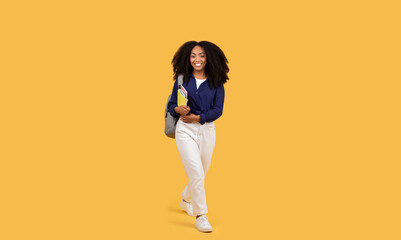 Fototapeta na wymiar Joyful black student with curly hair, carrying backpack and copybooks, strolls on yellow background