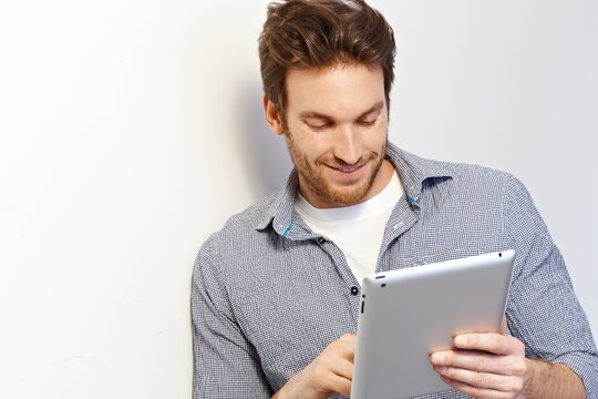 Happy white man working using tablet computer leaning against white wall. Copy space, casual clothing.
