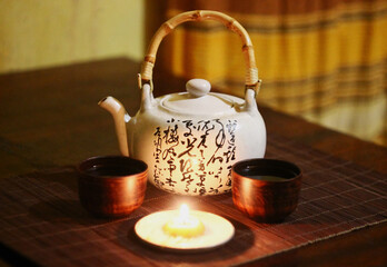 Chinese teapot with hieroglyphs, small clay tea cups in an ethnic atmosphere on a candlelit table.