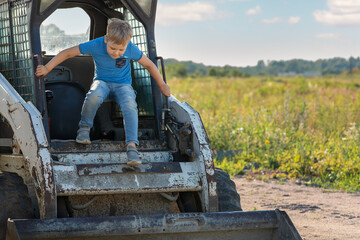 Cute boy in blue clothes coming out of bobcat cab. Rural meadow field background with free space for text