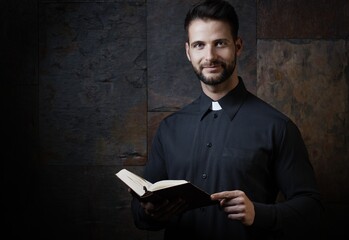 Portrait of handsome young catholic priest with prayer book against dark background.