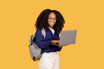 Cheerful young black lady with backpack and headphones holding laptop and smiling at camera over yellow background