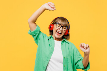Elderly blonde woman 50s years old she wear green shirt glasses casual clothes listen to music in headphones dance raise up hands isolated on plain yellow background studio portrait Lifestyle concept - Powered by Adobe