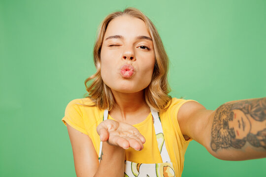 Close up young housewife housekeeper chef cook baker woman wears apron yellow t-shirt do selfie shot on mobile cell phone blow air kiss wink isolated on plain green background. Cooking food concept.