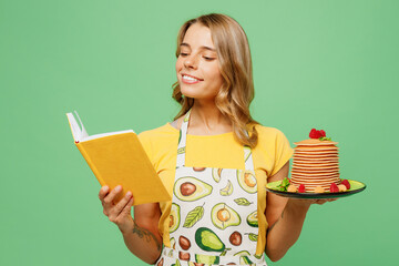Young fun housewife housekeeper chef cook baker woman wear apron yellow t-shirt hold in hand plate with pancakes read book recipe isolated on plain pastel green background studio Cooking food concept.