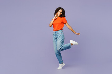 Full body side view little kid teen girl of African American ethnicity she wears orange t-shirt put hand on face raise up leg isolated on plain pastel purple background. Childhood lifestyle concept.