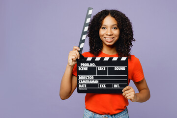Little kid teen girl of African American ethnicity in orange t-shirt hold in hand classic black film making clapperboard isolated on plain pastel purple background studio Childhood lifestyle concept