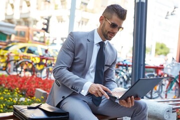 Businessman sitting on brench working with laptop on street.