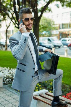 Portrait of young elegant businessman talking on smartphone on city street holding suitcase.