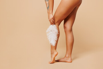 Full body close up cropped young lady woman with slim body perfect skin wearing nude top bra lingerie stand touch calf leg with feather isolated on plain beige background. Lifestyle diet fit concept.
