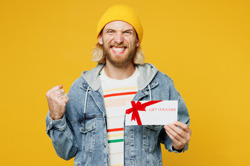 Young man wear denim shirt hoody beanie hat casual clothes hold gift certificate coupon voucher card for store do winner gesture isolated on plain yellow background studio portrait. Lifestyle concept