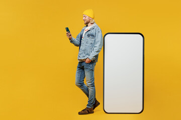 Full body side view young man wear denim shirt hoody beanie hat casual clothes big huge blank screen mobile cell phone with area using smartphone isolated on plain yellow background Lifestyle concept