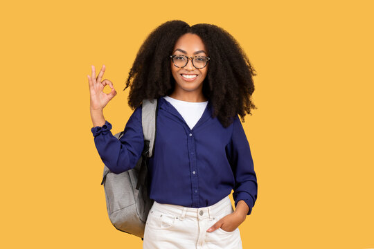 Happy black female student with backpack in glasses shows ok sign with hand isolated on yellow studio background