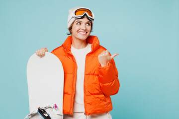 Young woman wear padded windbreaker jacket hat ski goggles mask hold snowboard point index finger aside on area travel rest spend weekend winter season in mountains isolated on plain blue background.