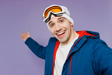 Close up skier man wear blue windbreaker jacket ski goggles mask hat do selfie shot mobile cell phone point aside spend extreme weekend winter season in mountains isolated on plain purple background.