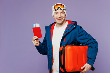 Traveler man wear blue windbreaker jacket ski goggles mask hat hold passport ticket bag isolated on plain purple background. Tourist travel abroad in free time rest getaway. Air flight trip concept.