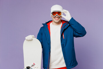 Smiling happy cool fun man wear warm blue windbreaker jacket touch ski goggles mask hat hold snowboard look camera spend extreme weekend winter season in mountains isolated on plain purple background.