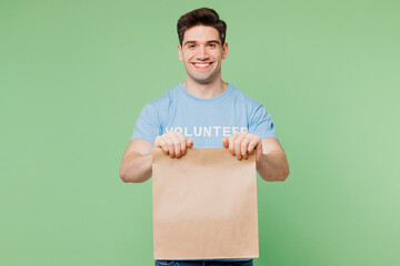 Young fun man wears blue t-shirt white title volunteer hold in hand paper brown blank bag delivery isolated on plain pastel green background. Voluntary free work assistance help charity grace concept.