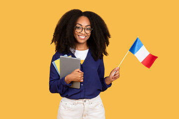 Excited african american lady student with copybooks, holding flag of France, posing on yellow...