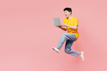 Full body young IT man he wearing yellow t-shirt casual clothes jump high hold use work on laptop pc computer isolated on plain pastel light pink color background studio portrait. Lifestyle concept.