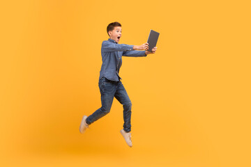Surprised teenage boy jumping with digital tablet in hands over yellow background