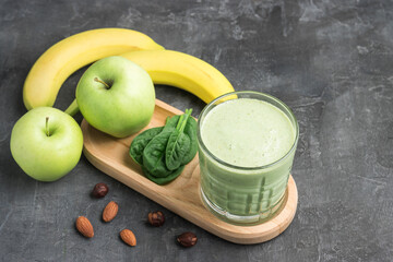 Green detox smoothie in a glass, blended vegetarian drink from spinach leaves, apple, banana fruit and nuts on dark background, healthy eating concept