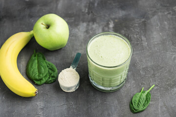 Green detox smoothie in a glass, measuring spoon with protein powder, blended vegetarian drink from spinach leaves, apple and banana fruit on dark background, healthy eating concept