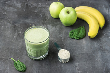 Green detox smoothie in a glass, measuring spoon with protein powder, blended vegetarian drink from spinach leaves, apple and banana fruit on dark background, healthy eating concept