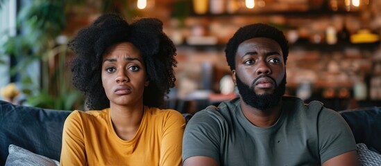 Concerned young African American couple watching TV, displaying skepticism and anxiety, upset due to a problem. - Powered by Adobe