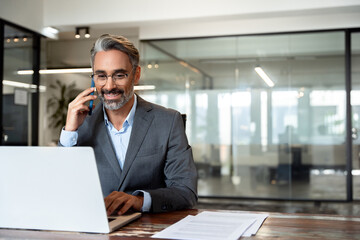 Middle aged Latin or Indian businessman having call on smartphone with business partners or clients. Smiling mature Hispanic man sitting at table talking by mobile cellphone at workplace in office. 