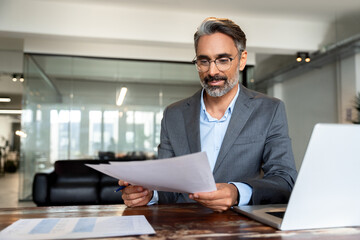 Smiling Latin or Indian male business man accountant analyst holding documents, work at laptop computer doing online trade market tech research. Focused Hispanic businessman with paperwork in office.