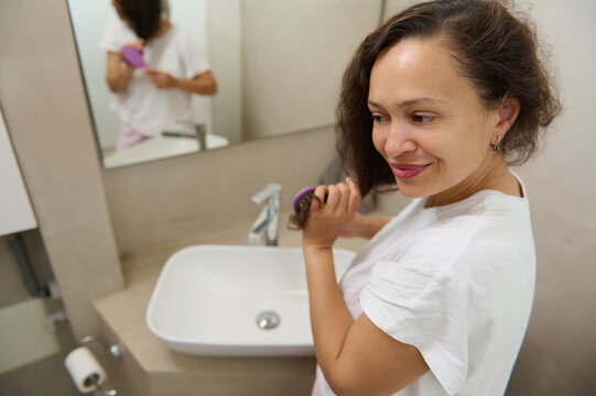 Beautiful young woman brushing hair with hair comb, smiling enjoying morning beauty procedures in the home bathroom